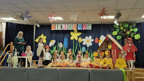 Zdjęcie przedstawia przebrane dzieci siedzące i stojące na scenia występujące przed innymi dziećmi