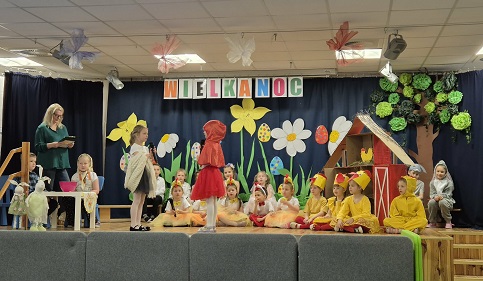 Zdjęcie przedstawia dwoje przebranych dzieci stojących na scenie rozmawiających ze sobą za nimi siedzi reszta dzieci biorących udział w uroczystości