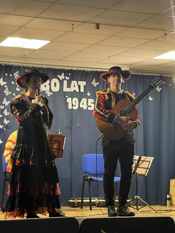 Obraz przedstawia muzyków na scenie występujących przed dziećmi.