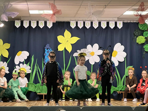 Zdjęcie przedstawia trzy dziewczynki stojace na scenie za nimi inne siedzące dzieci