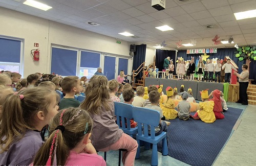 Zdjęcie przedstawia występującą na scenie grupę dzieci przed nimi siedząca widownia