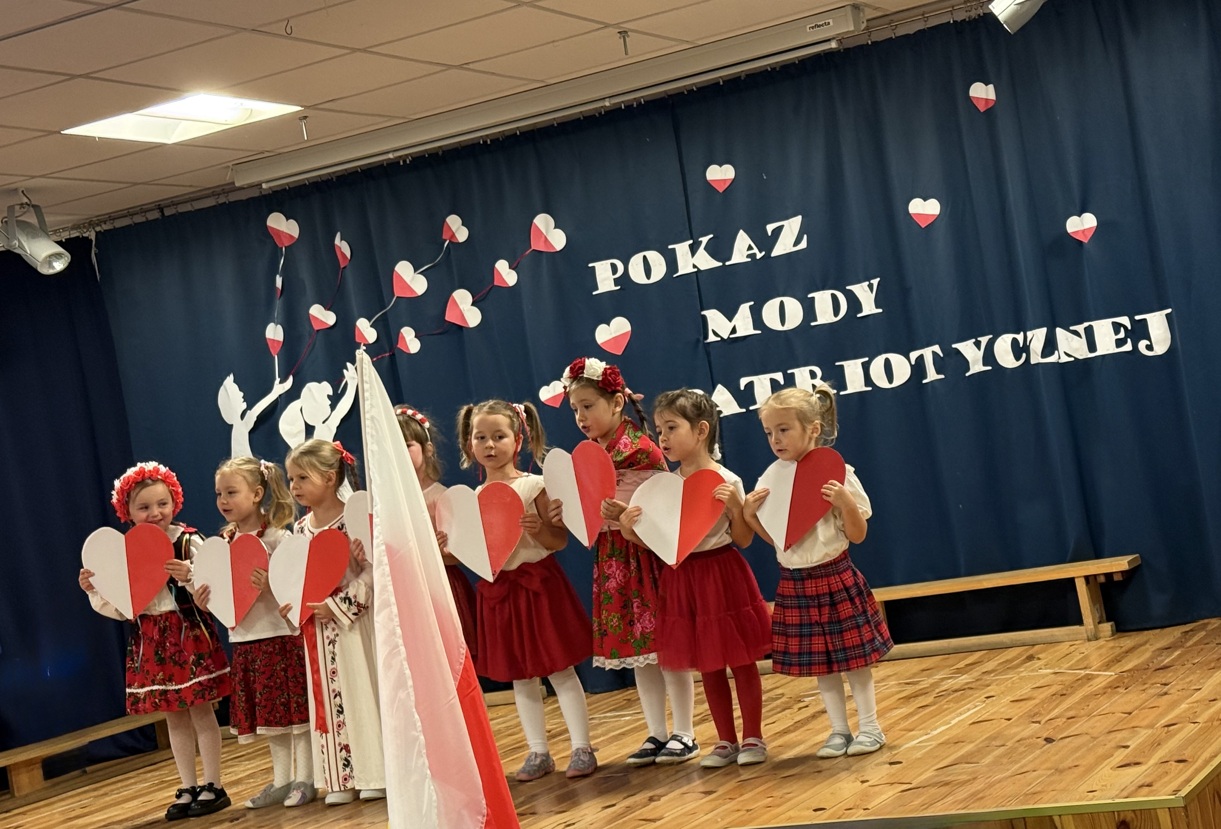Zdjęcie przedstawia grupę dzieci na scenie trzymającą biało-czerwone serca w dłoniach