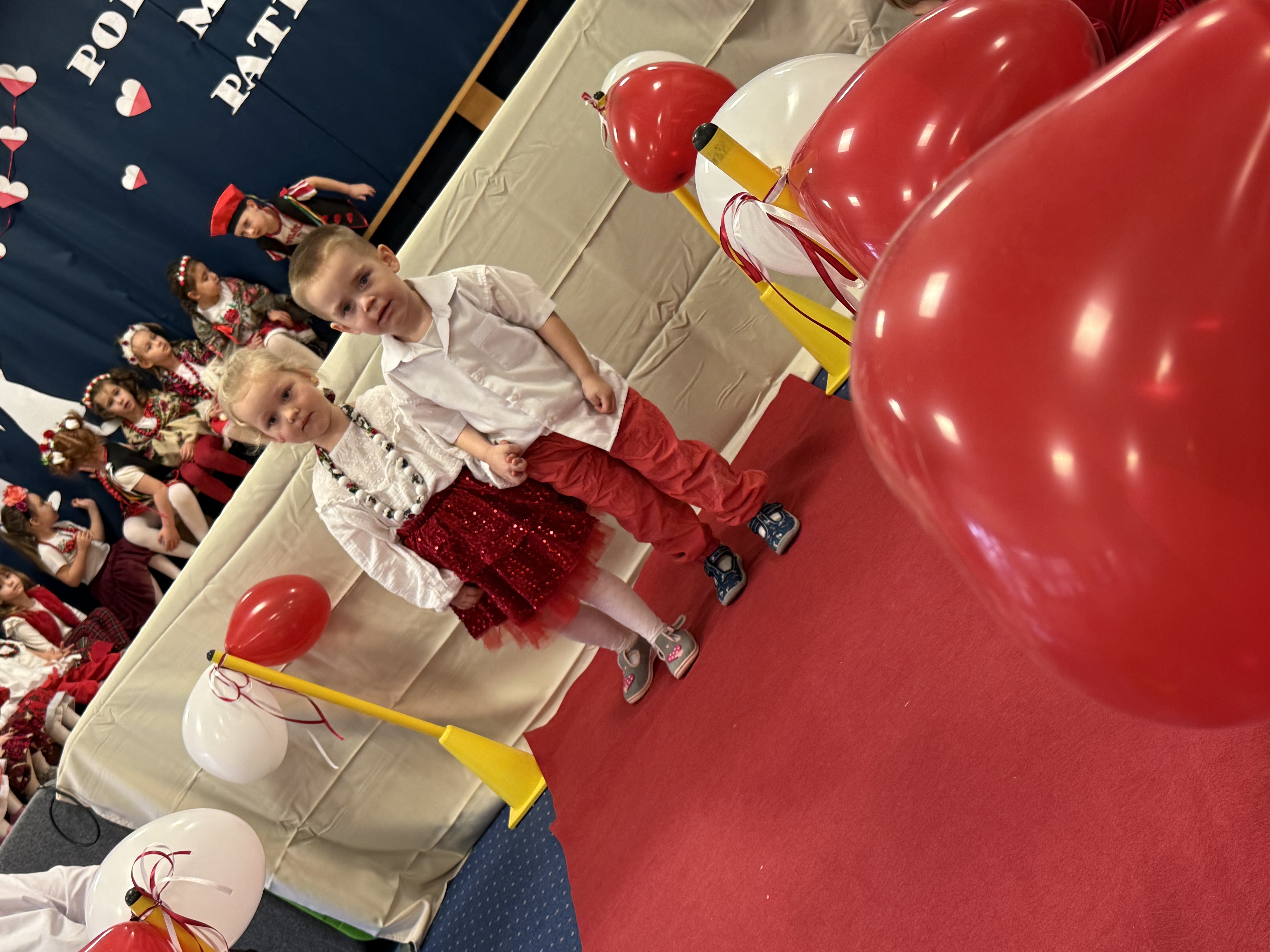 Zdjęcie przedstawia dziewczynkę i chłopca podczas pokazu mody, w tle grupa dzieci na scenie