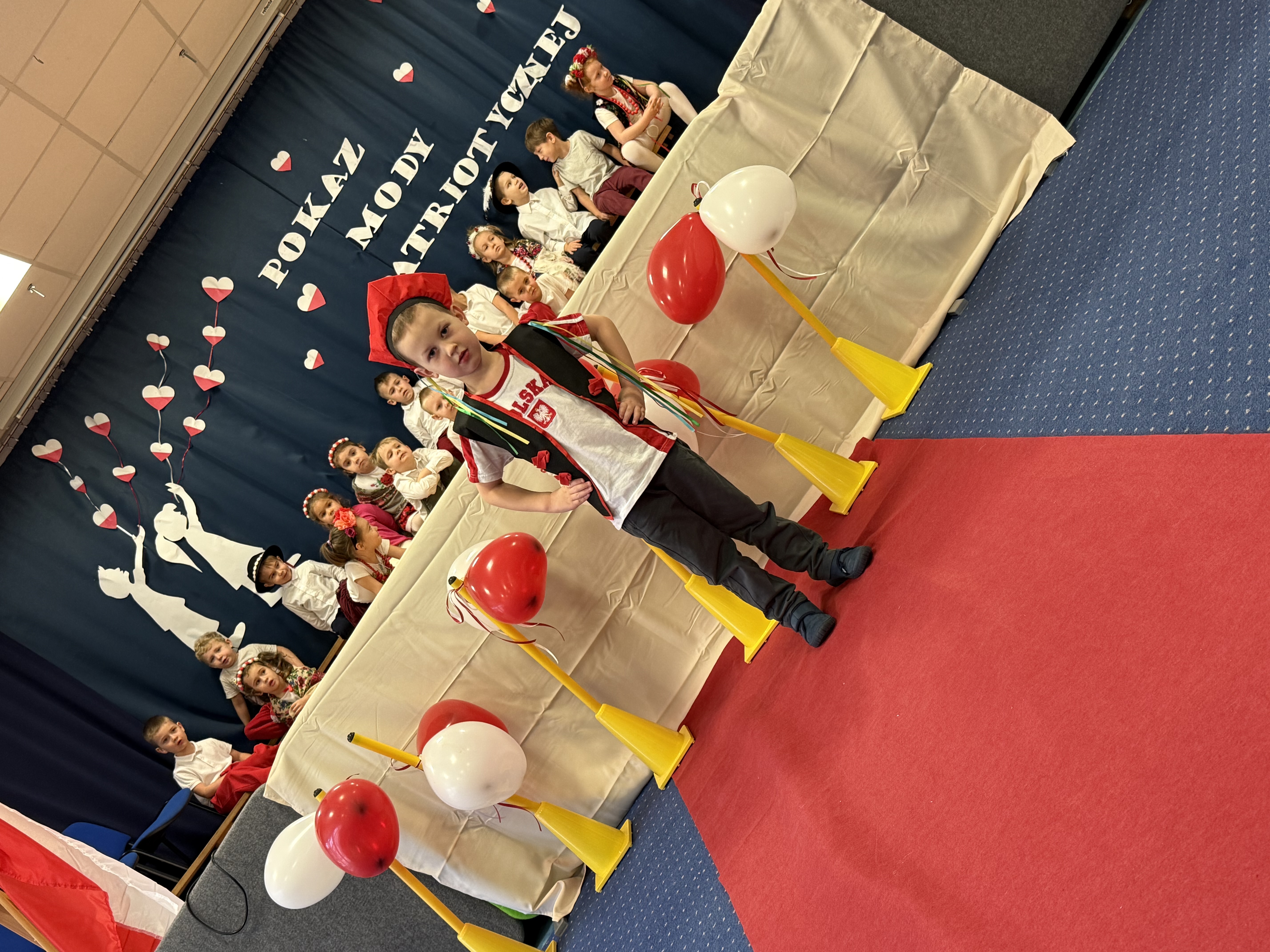 Zdjęcie przedstawia chłopca podczas pokazu mody, w tle grupa dzieci na scenie