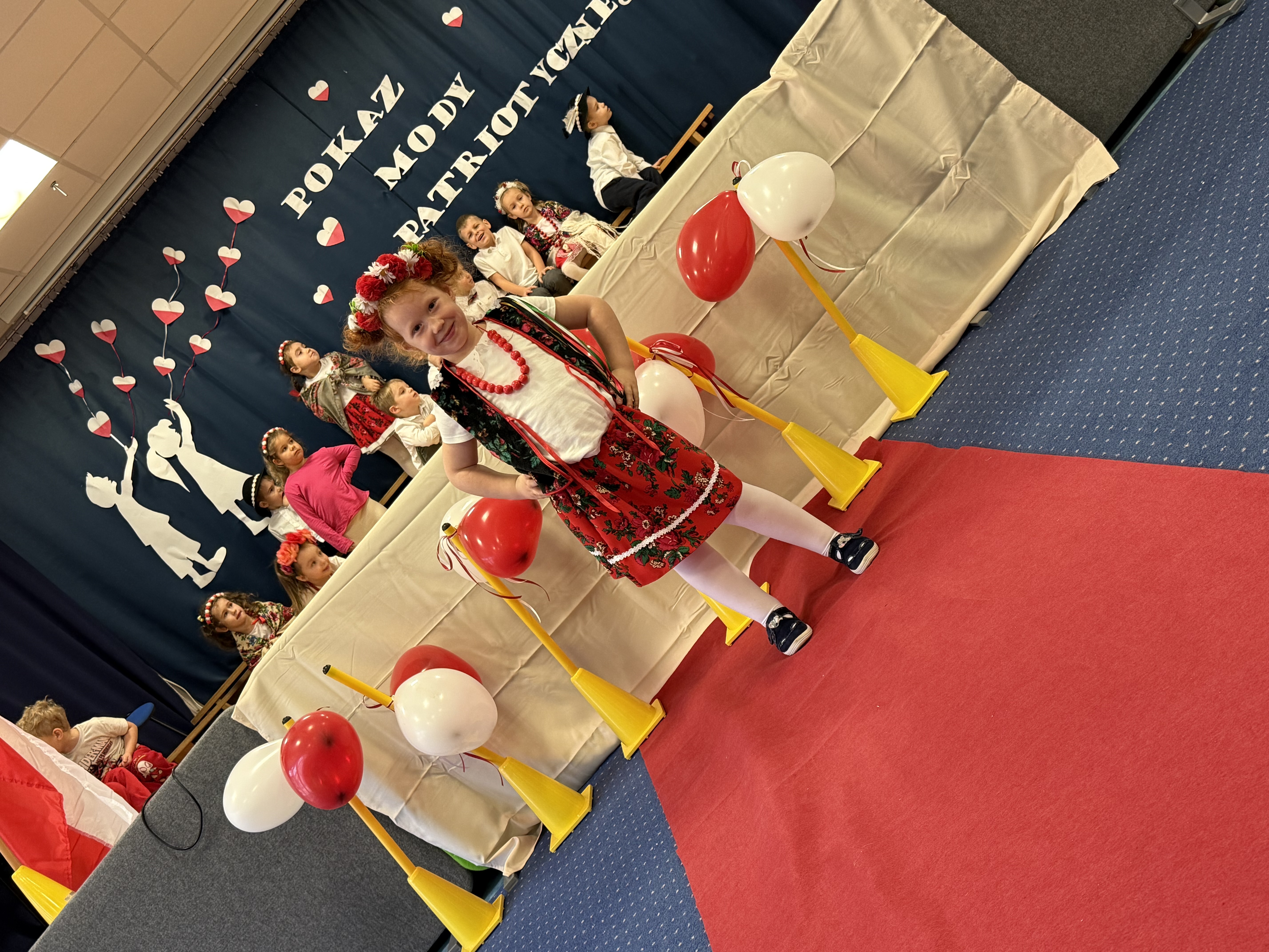 Zdjęcie przedstawia dziewczynkę podczas pokazu mody, w tle grupa dzieci na scenie