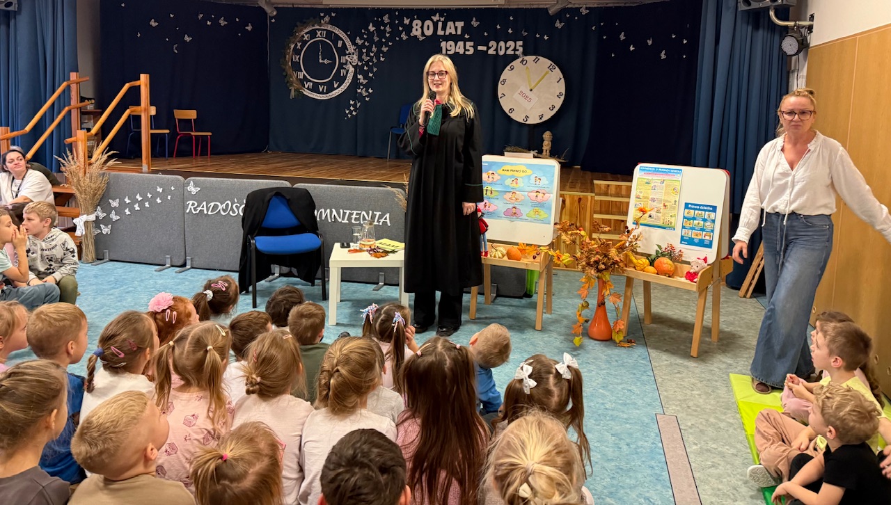 Zdjęcie przedstawia dwie osoby dorosłej na tle sceny oraz dzieci na widowni