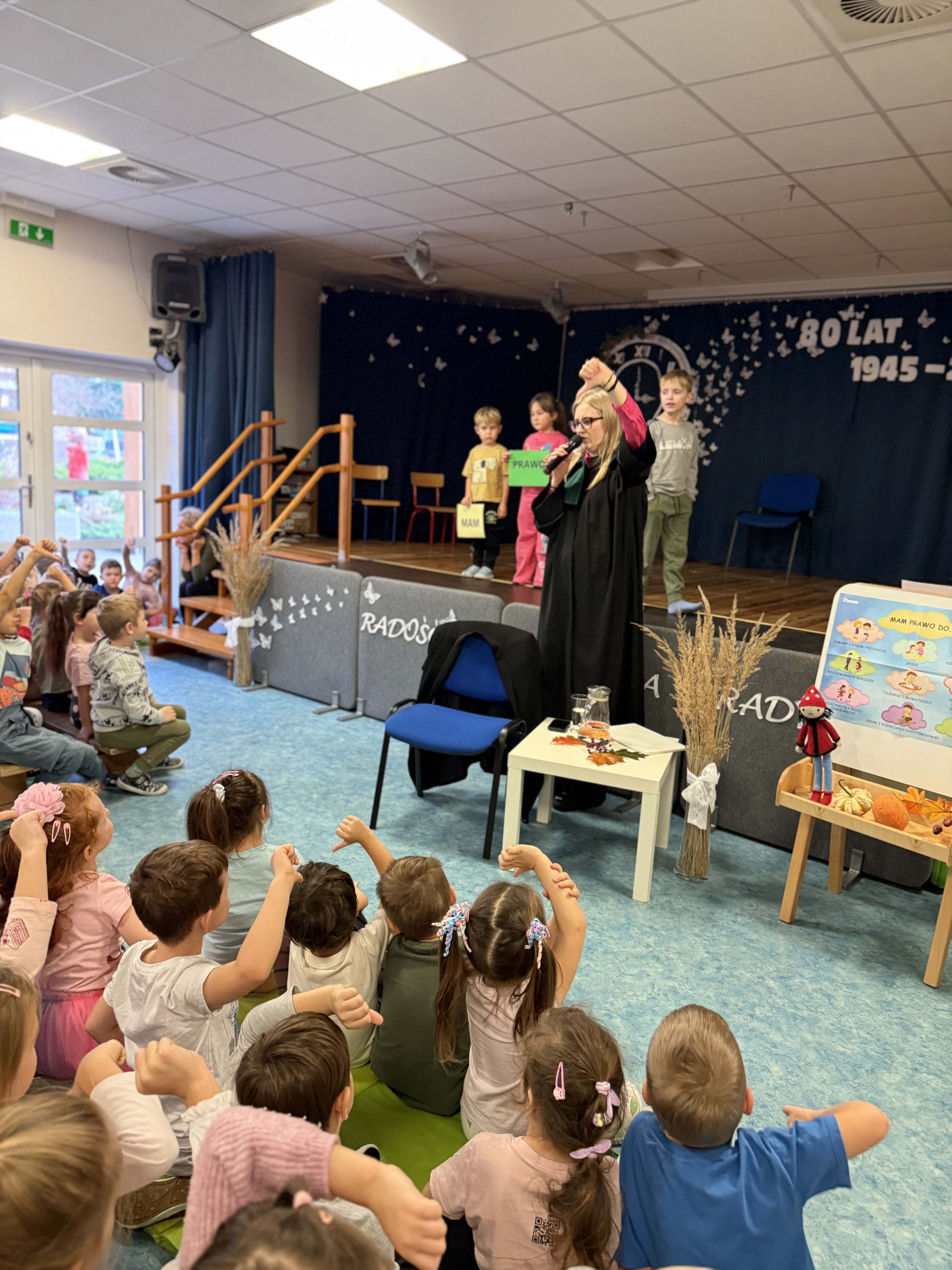 Zdjęcie przedstawia grupę dzieci na scenie, widowni oraz kobietę pokazującą kciukiem w dół