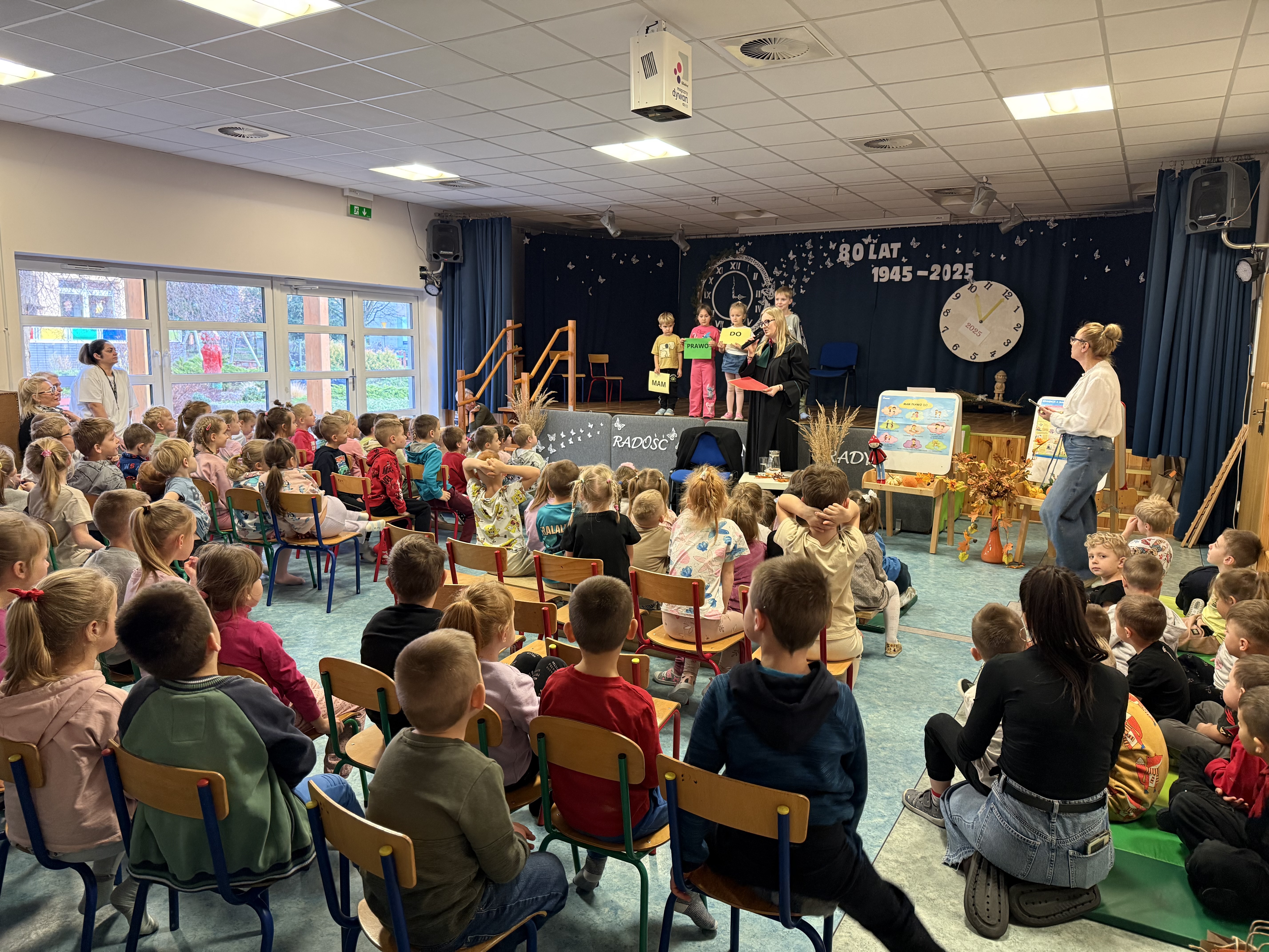 Zdjęcie przedstawia grupę dzieci oraz dorosłych w sali sportowo-widowiskowej przedszkola