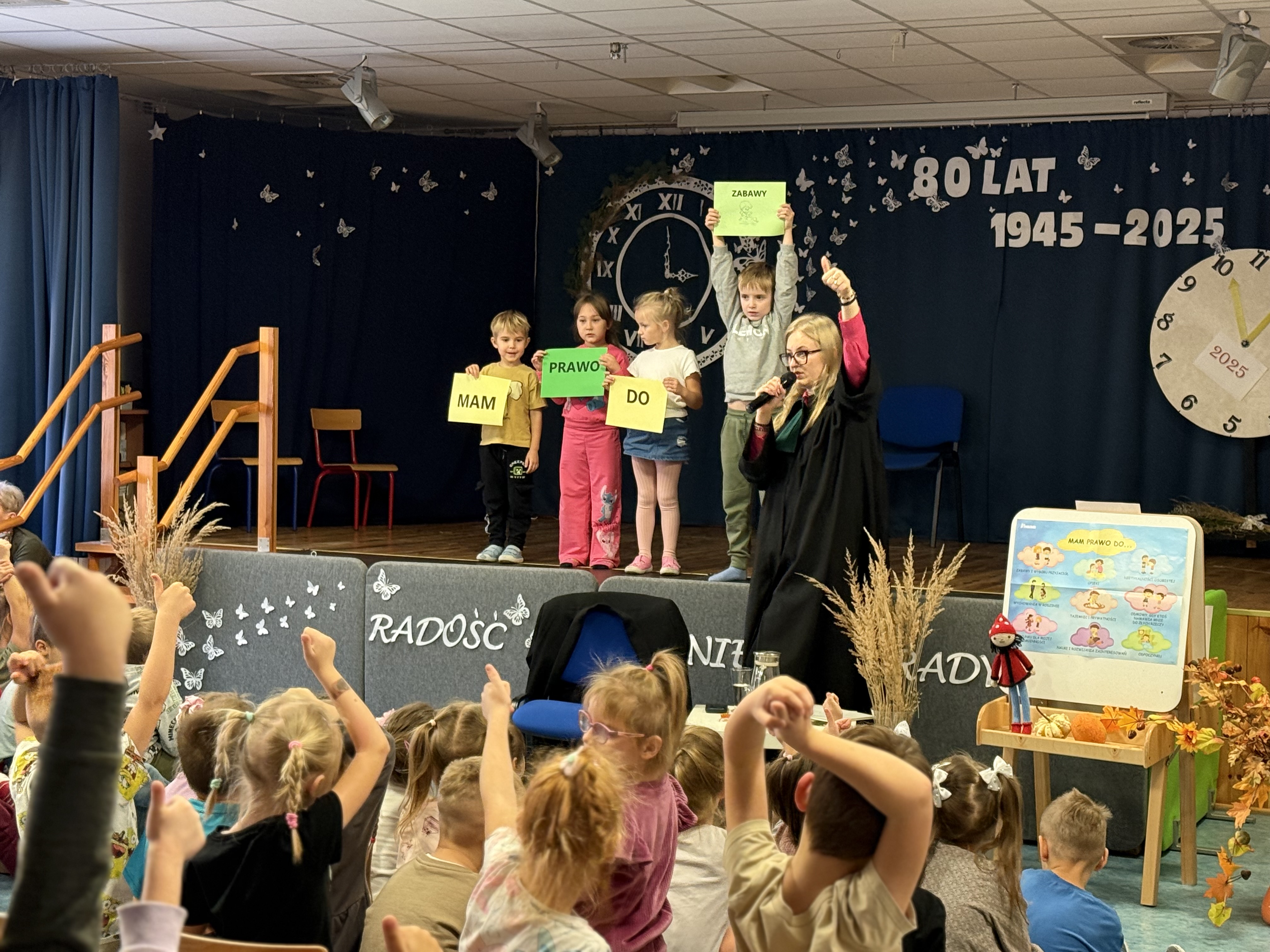 Zdjęcie przedstawia grupę dzieci na scenie oraz adwokat a ponad to dzieci na widowni