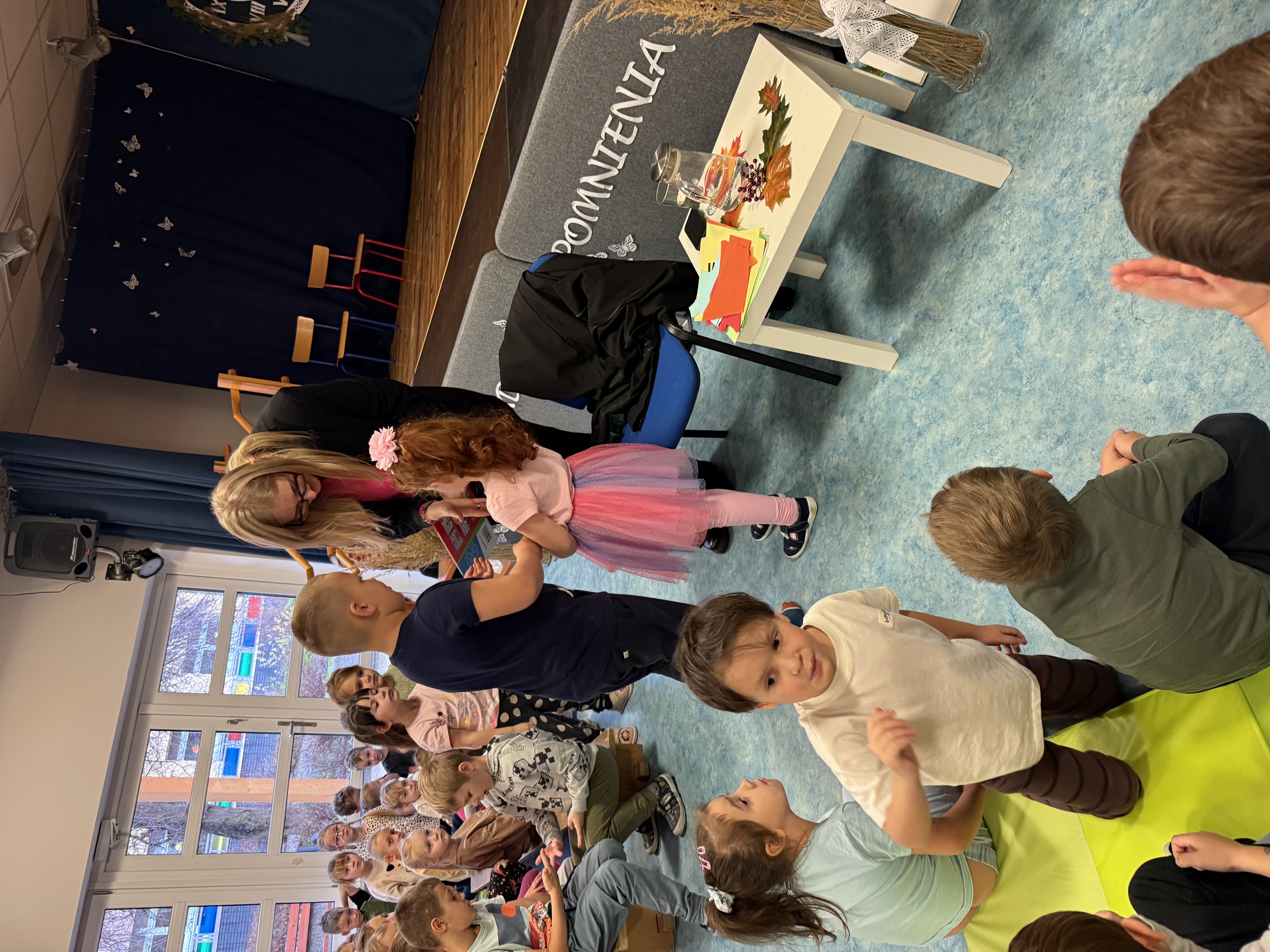 Zdjęcie przedstawia dziewczynkę oraz chłopca wręczających upominek kobiecie, w tle grupa dzieci